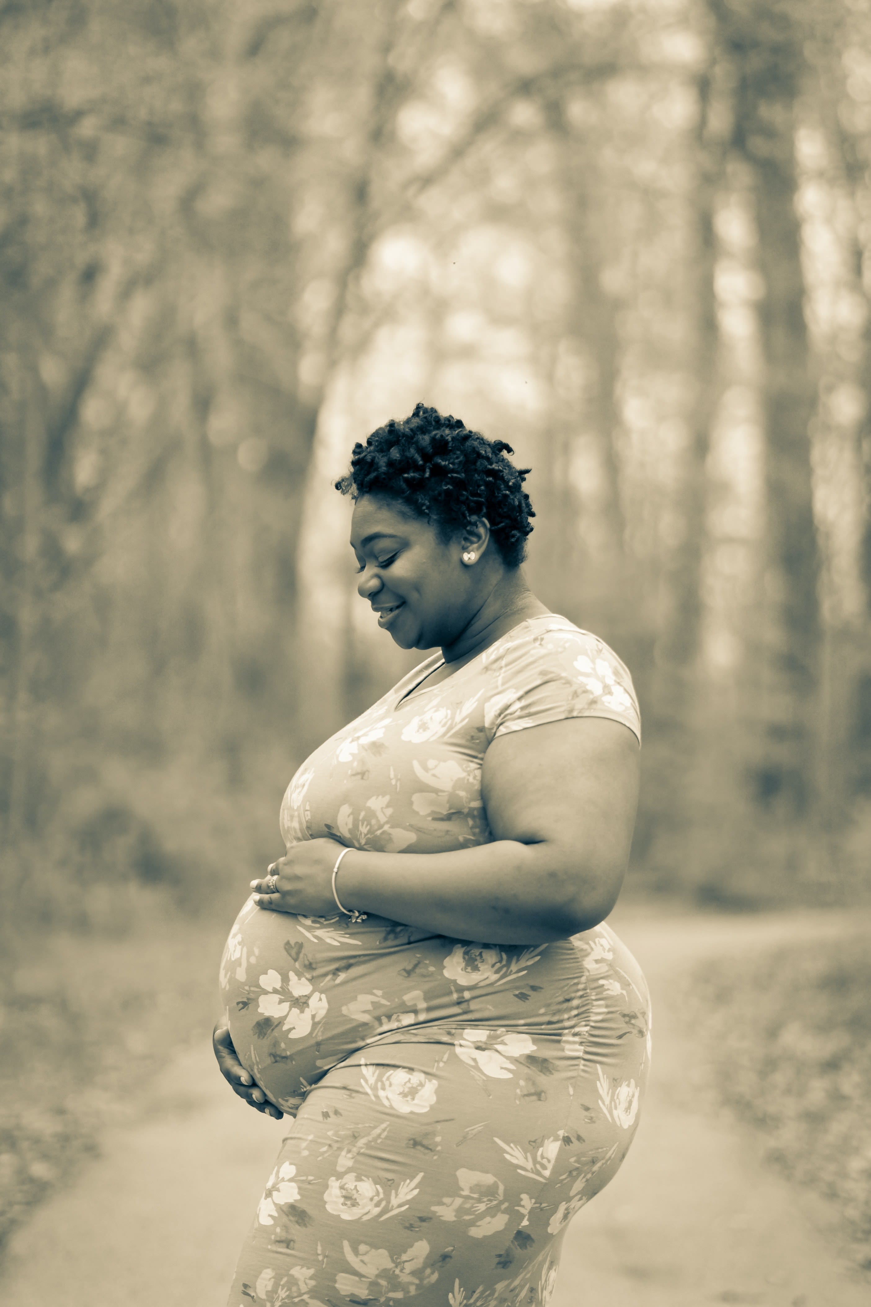 Photo of a pregnant woman, wearing a flowery dress, standing in the woods..