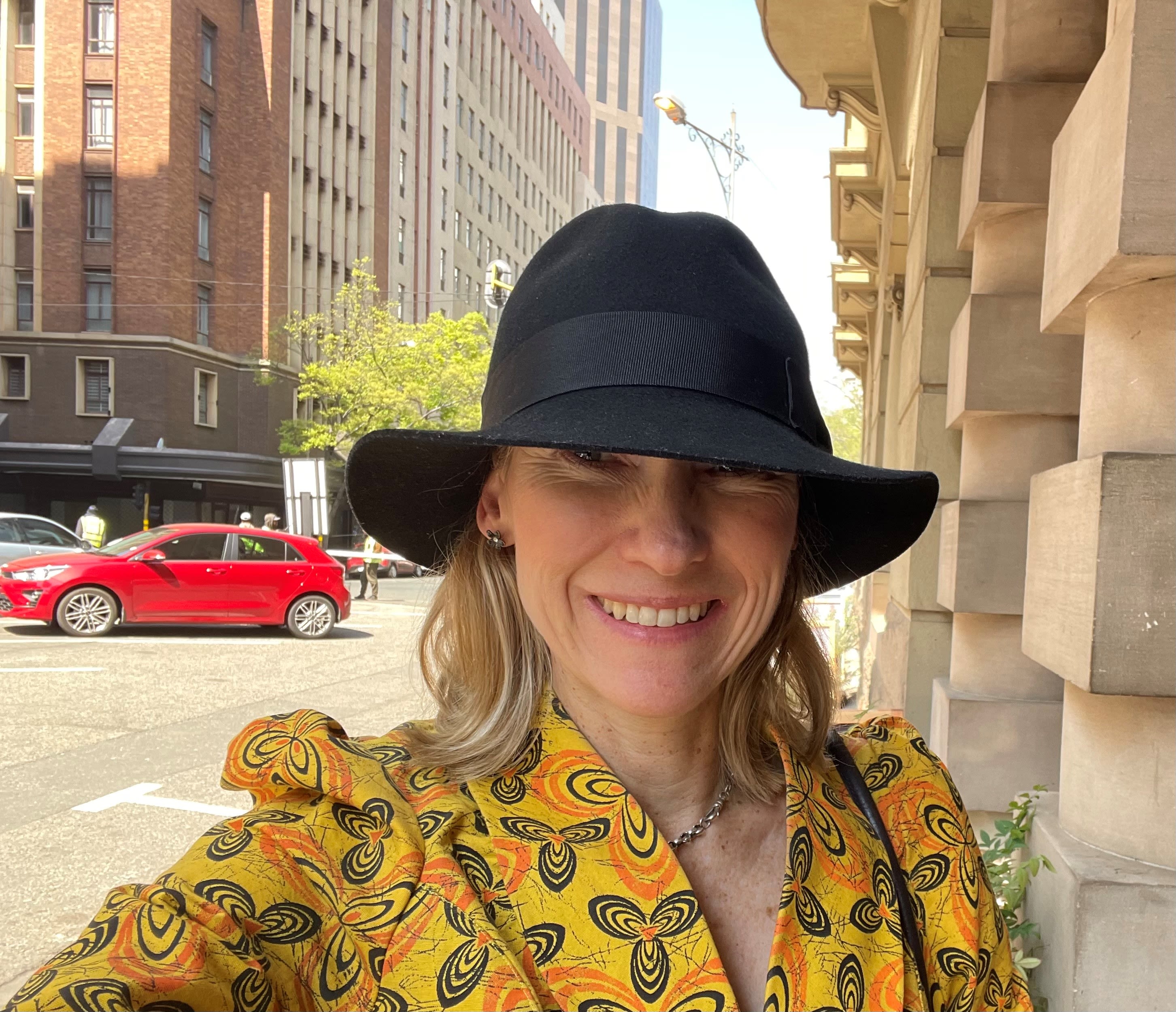 Blonde woman, smiling, wearing a yellow patterned jacket and a black hat.