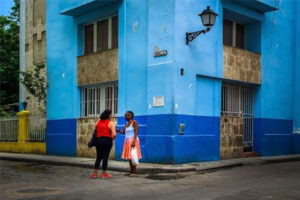 Two women speaking to each other in front of a bright blue building in a Cuban street