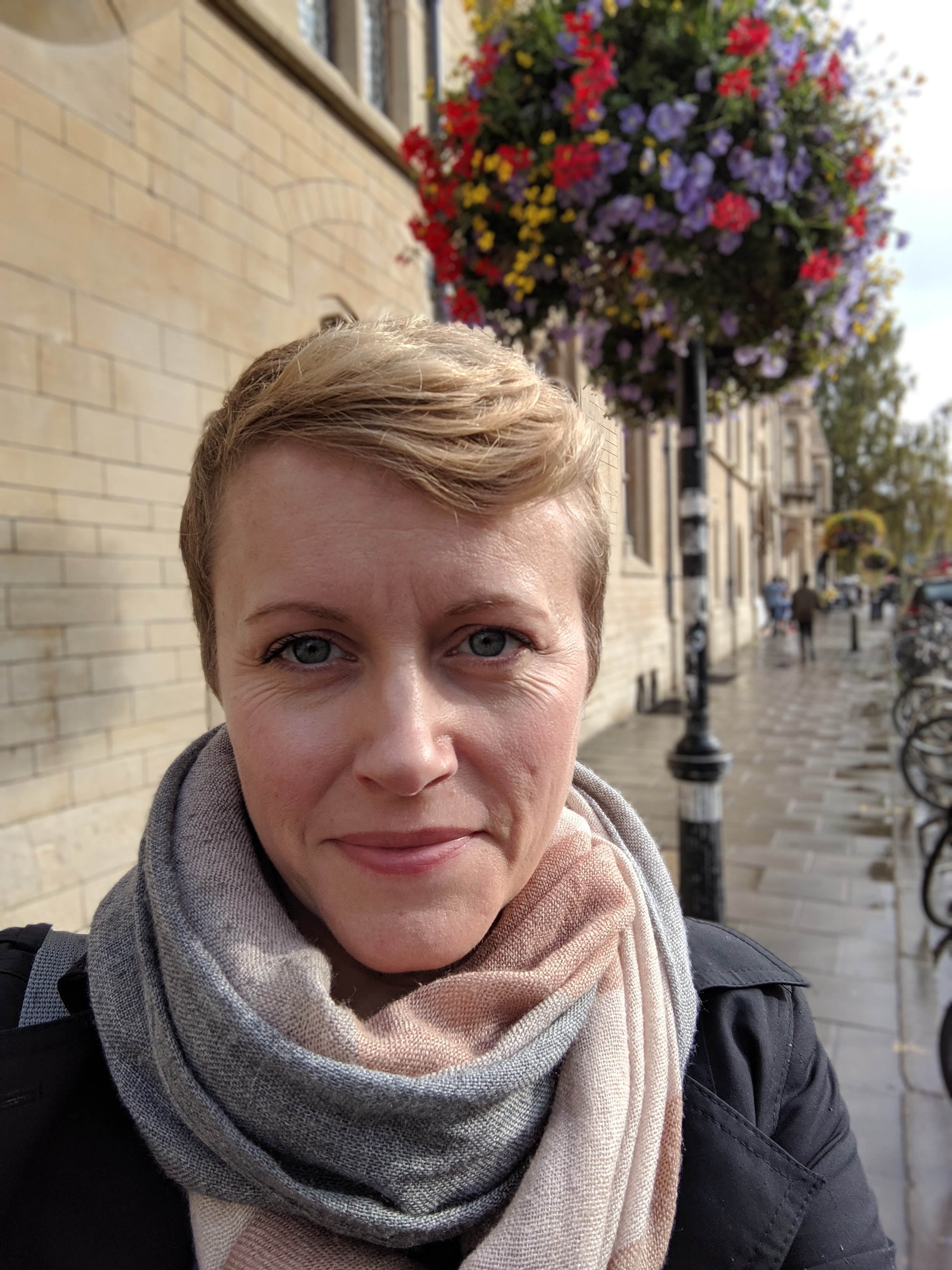 Picture of a white woman smiling with a grey and beige scarf, short dark blonde hair
