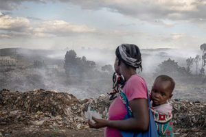 Woman carrying baby on back looks over a rubbish dump and air pollution
