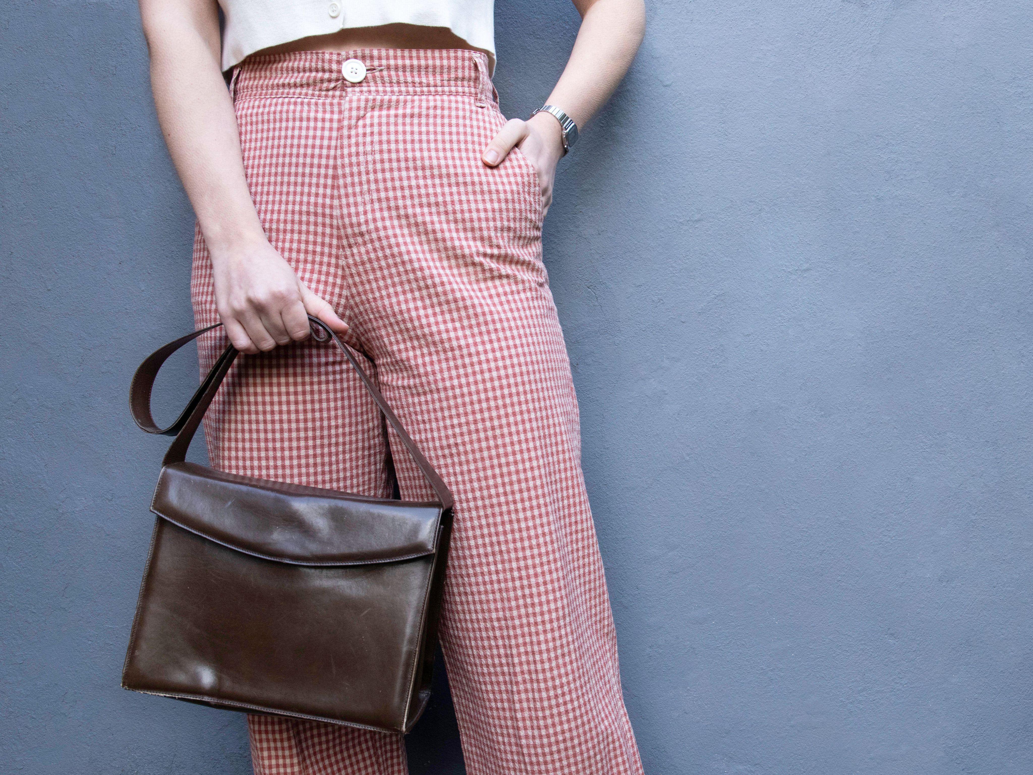 Bottom half of person wearing red gingham trousers and holding vintage leather bag