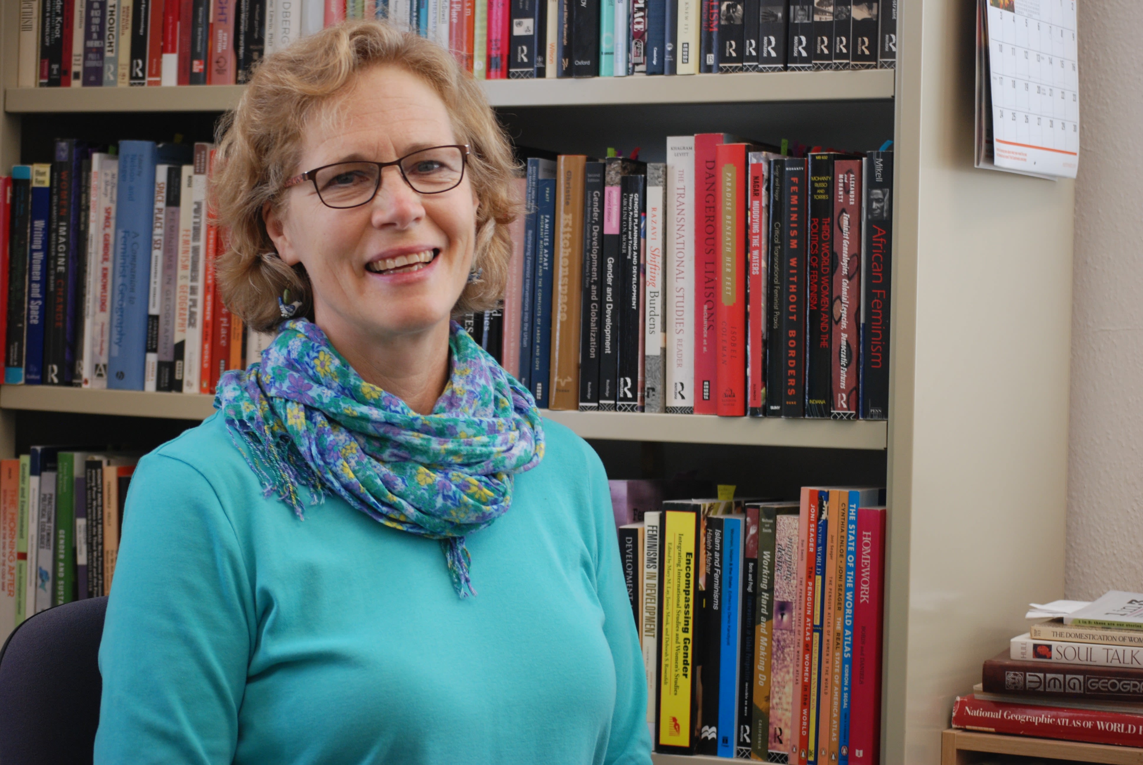 Ann has medium length curly blonde hair. A teal blue top and a blue and yellow scarf with small flowers. Ann also wears glasses with dark thin frames.