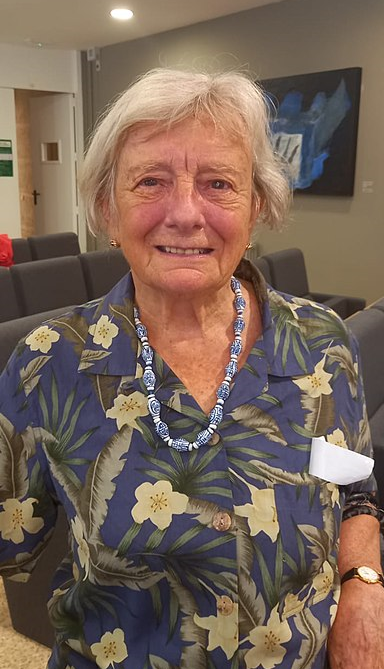 Picture of Janet Momsen, she has short gray hair and a colorful blue shirt with a tropical plant design. She is also wearing a striking white and blue beaded necklace.
