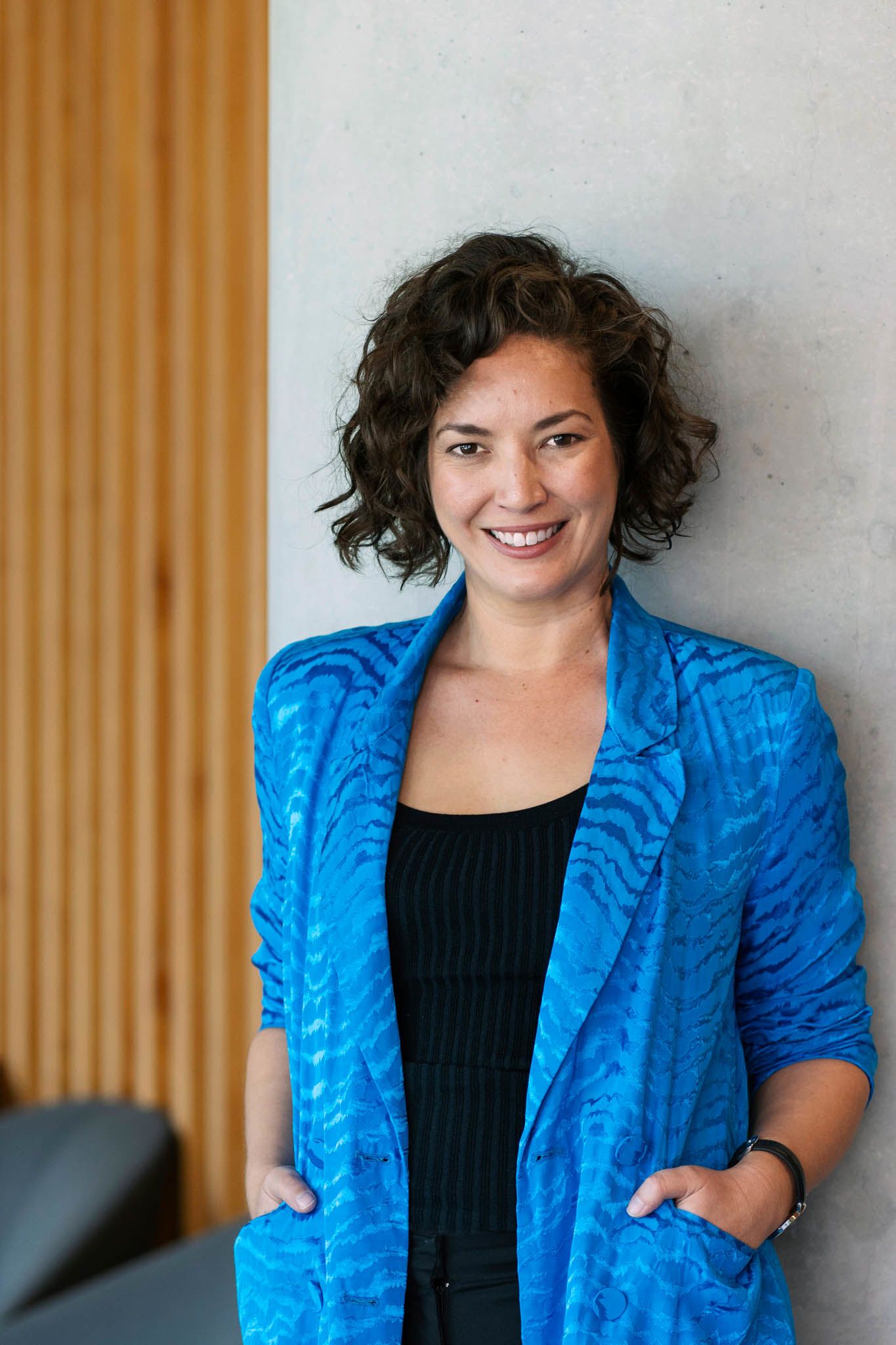 Image of Alana Kamp. She has a black top, a very bright blue shiny blazer, a curly brown bob haircut, and a big smile. She has her hands in her pockets.
