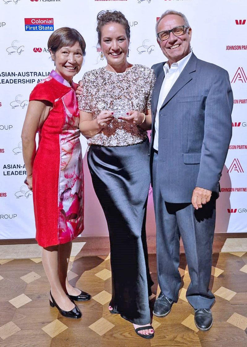 Alanna has a long black silk skirt, her mother (left) is wearing a bright red cocktail dress, her father (right) is wearing a gray suit. This is taken at an awards ceremony called the 2024 Asian Australian Leadership Award.