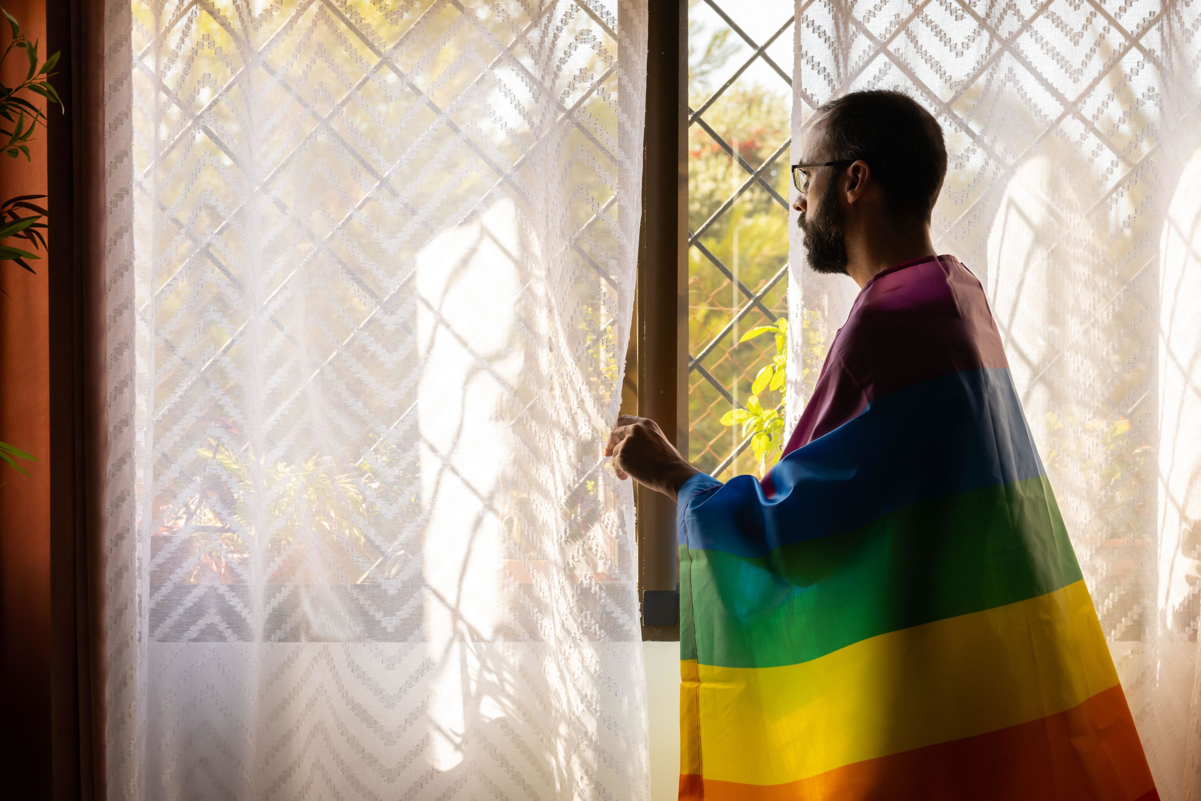 Man with rainbow flag on shoulders looking out window outdoors
