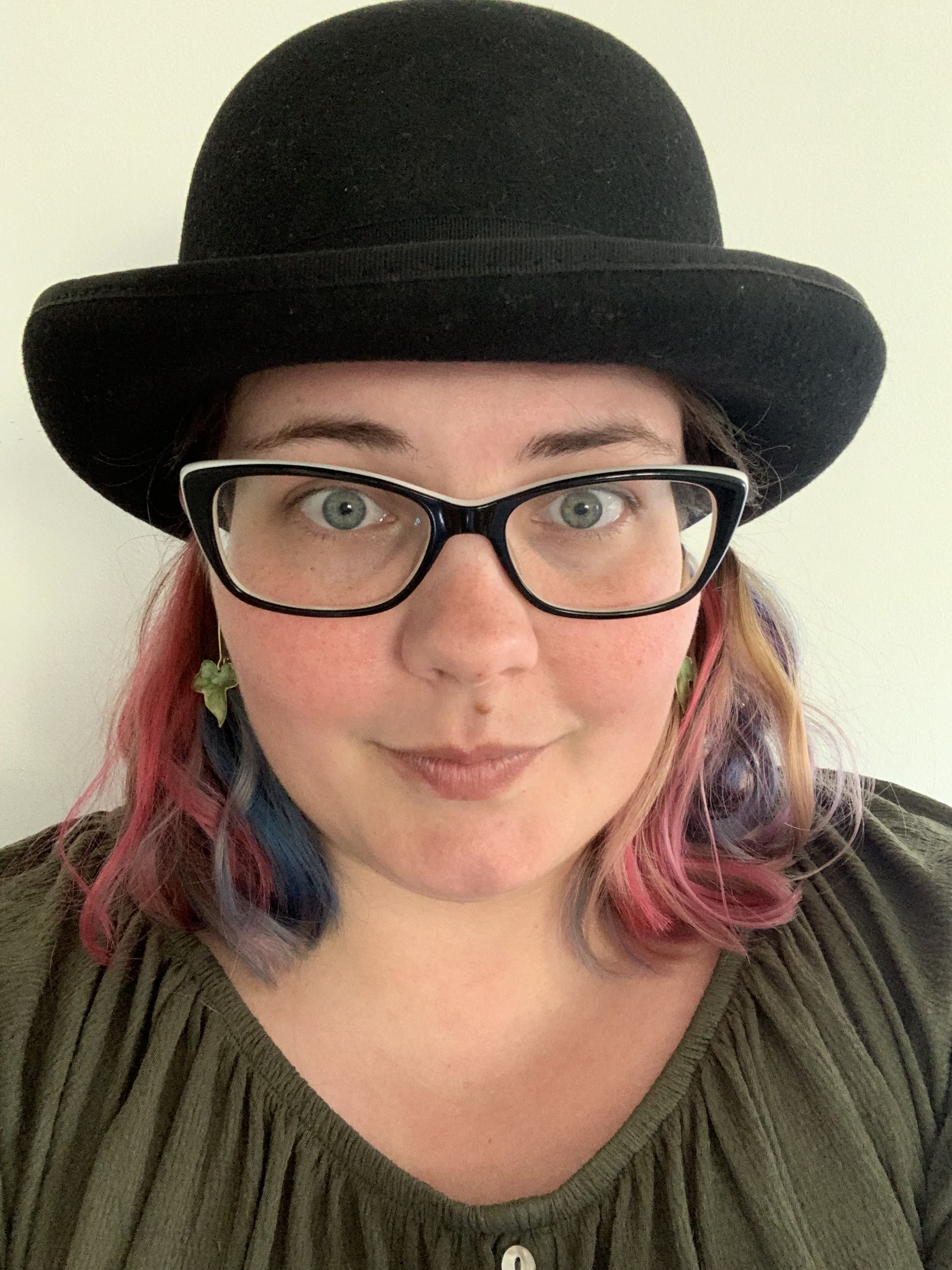 A young woman with pink and blue hair, wearing a black bowler hat, glasses and an olive green shirt