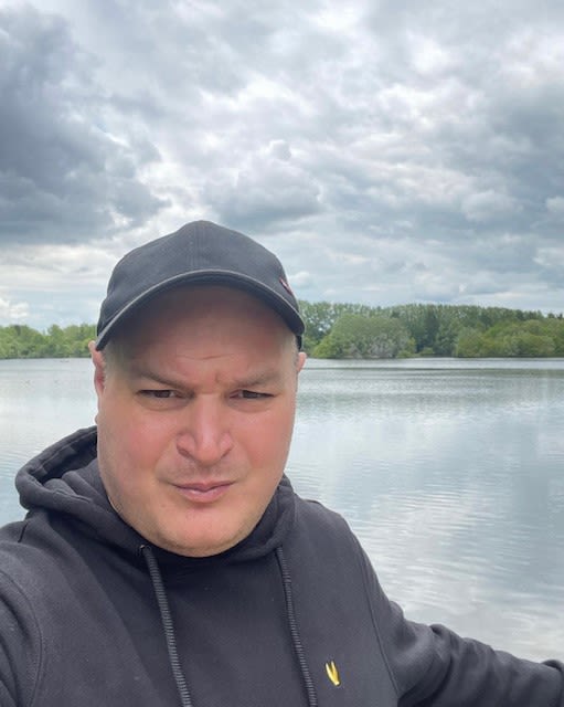 Photo of a man with a baseball cap and grey sweatshirt, sitting in a boat on a lake.