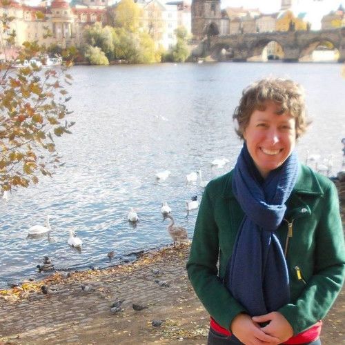 Image of a woman with short curly blonde hair, a bright blue scarf and a green jacket. She's standing at a pond with several waterbirds in it.