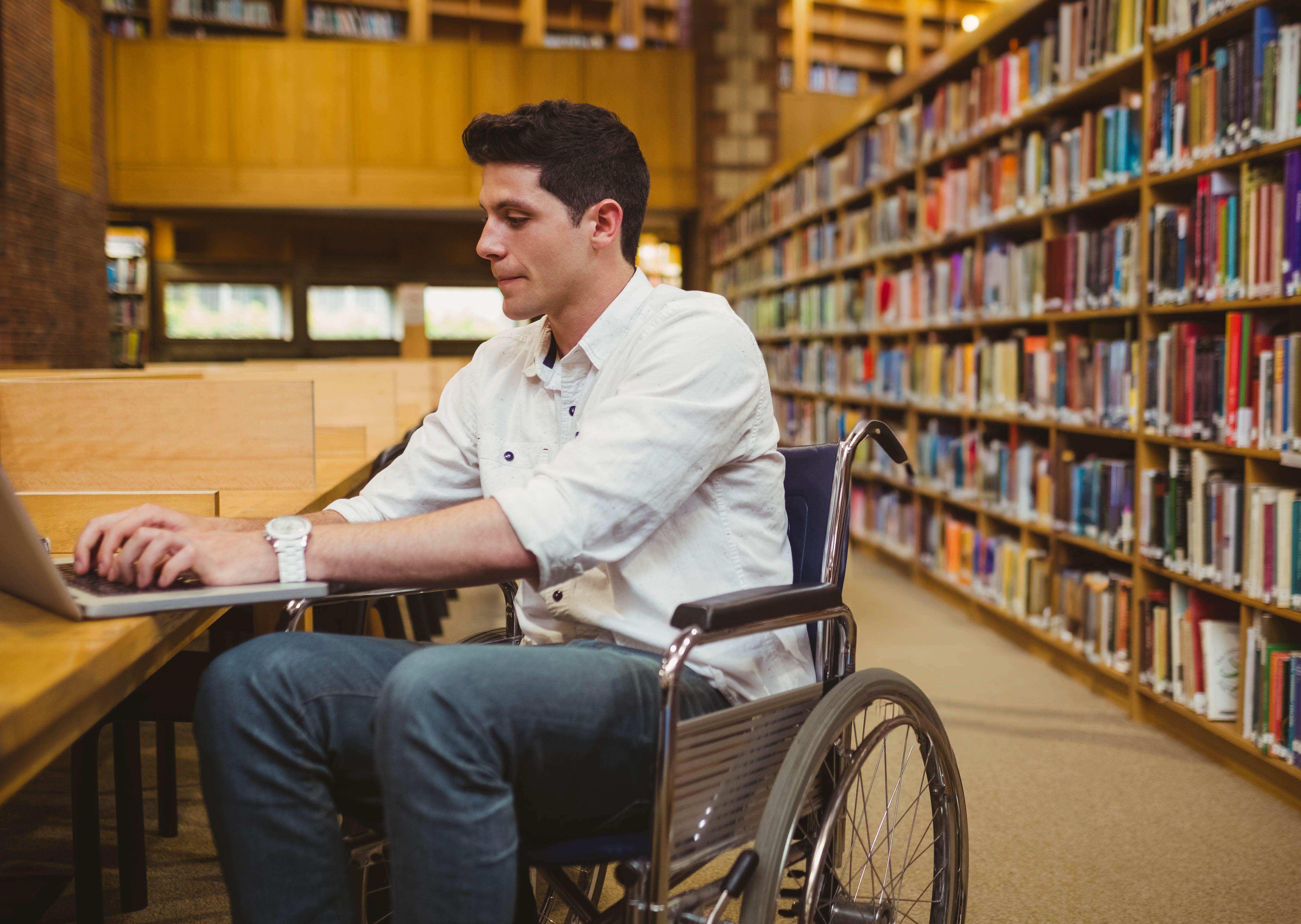 Man in wheelchair typing on laptop at wooden study table inside library with bookshelves.